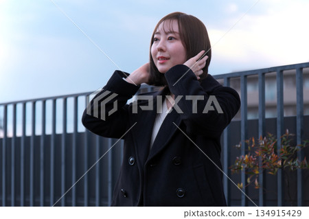 冬日傍晚，一位身穿黑色外套的女子在戶外迎著風整理頭髮，這是一幅肖像畫。 134915429