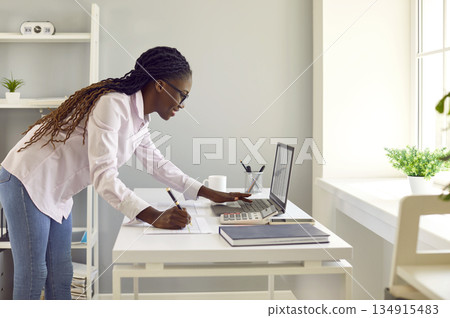 African American woman who works as accountant or secretary uses laptop and takes notes 134915483