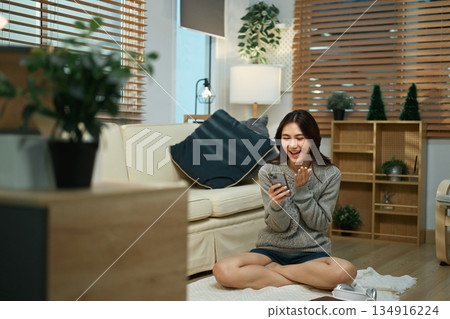 Young woman smiles while using a phone at home, sitting on the floor in a cozy living room during relaxed 134916224