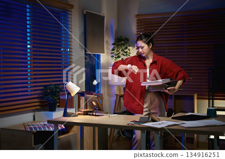Interior designer checks her watch while holding material samples during late night work in a home office Interior designer checks her watch while holding material samples during late night work in a home office 134916251