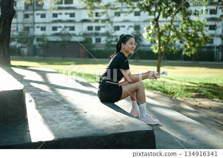 Smiling woman resting and listening to music during outdoor workout break in park Smiling woman resting and listening to music during outdoor workout break in park 134916341
