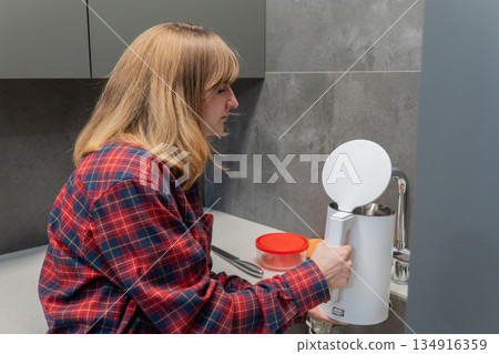 Caucasian woman filling electric kettle in kitchen wearing plaid shirt pouring water into white kettle near red canister gray cabinets focused on countertop calm morning routine domestic beverage 134916359