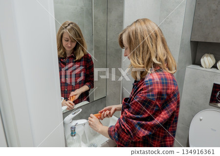 Woman washing and wiping bathroom sink with soap and cloth, focused hand care and surface sanitizing, plaid shirt visible, faucet and mirror details, compact modern bathroom, practical evening tidyup 134916361