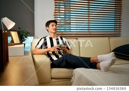 Man relaxes on his sofa with a warm cup of coffee, enjoying a quiet day off and letting his thoughts wander freely 134916369