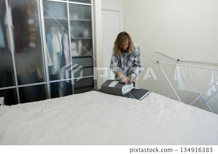Female neatly irons sheet with intent focus, Woman meticulously presses her bed linens with precision, An individual diligently smooths bedding using compact iron board with careful attention Female neatly irons sheet with intent focus, Woman meticulously presses her bed linens with precision, An individual diligently smooths bedding using compact iron board with careful attention 134916383