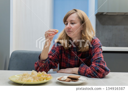 white woman snacking at kitchen table, tastetesting chips and cookies, thoughtful expression, plaid shirt, home interior, casual afternoon break, sampling flavors for snack blog review 134916387