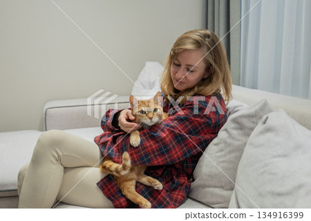 Comfortable woman with feline friend, Modern apartment with woman embracing her orange feline pet warmly, Relaxed female individual sharing cozy moment with her beloved pet cat indoors Comfortable woman with feline friend, Modern apartment with woman embracing her orange feline pet warmly, Relaxed female individual sharing cozy moment with her beloved pet cat indoors 134916399