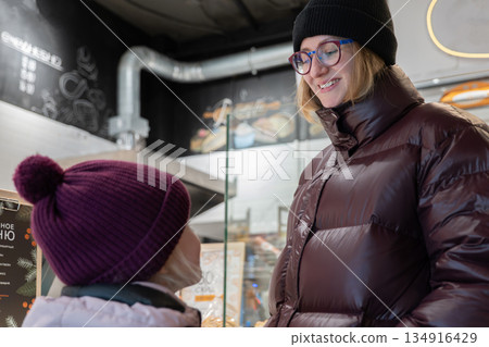 Caucasian woman smiling towards child, genuine warmth and eye contact, maroon puffer jacket and knit hat, bakery lights and pastry display creating intimate cozy vibe, capturing tender parenting Caucasian woman smiling towards child, genuine warmth and eye contact, maroon puffer jacket and knit hat, bakery lights and pastry display creating intimate cozy vibe, capturing tender parenting 134916429