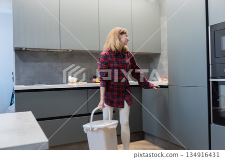 Blonde white woman standing with recycling bin in kitchen, pausing in thoughtful pose while organizing waste. Casual homewear and minimalist interior create quiet domestic mood. Scene suggests Blonde white woman standing with recycling bin in kitchen, pausing in thoughtful pose while organizing waste. Casual homewear and minimalist interior create quiet domestic mood. Scene suggests 134916431