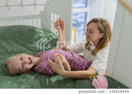 white mother tickling daughter on bed playful bedtime storyteller scene in kids room sunlight through window green patterned duvet giggling child relaxed casual clothes intimate mother daughter white mother tickling daughter on bed playful bedtime storyteller scene in kids room sunlight through window green patterned duvet giggling child relaxed casual clothes intimate mother daughter 134916444