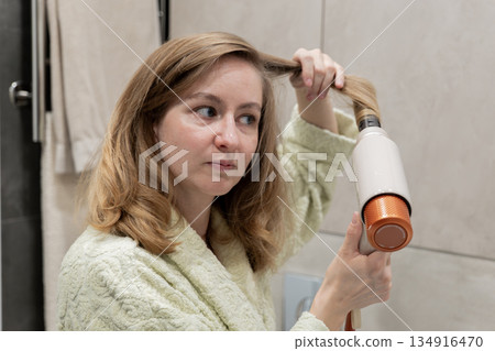 Closeup woman drying hair ends with hairdryer and round brush, focused expression and warm studio lighting, modern tiled background, content creator vibe demonstrating technique, intimate evening 134916470