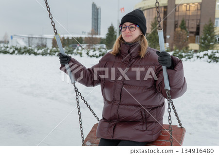 Closeup caucasian woman smiling on swing in winter wearing maroon puffer, black beanie and glasses, soft light, textured fabric details, candid fashion blogger vibe, urban plaza and apartment Closeup caucasian woman smiling on swing in winter wearing maroon puffer, black beanie and glasses, soft light, textured fabric details, candid fashion blogger vibe, urban plaza and apartment 134916480