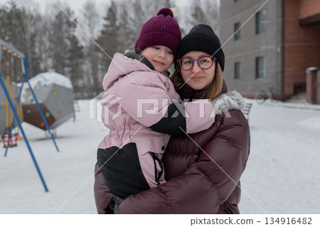 white nanny cradling daughter close during winter portrait, closeup of smiling faces, adult wearing glasses and black beanie, child in magenta hat and pink coat, soft skin contact and warm eye 134916482