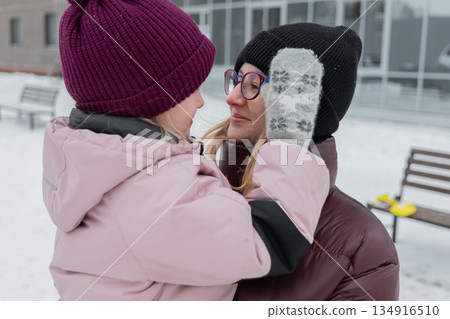 Closeup winter hug between grandmother and grandchild, mitten pressed to cheek, smiling adult wearing glasses and knit beanie, heavy puffer coats, soft snowfall, blurred urban background, intimate Closeup winter hug between grandmother and grandchild, mitten pressed to cheek, smiling adult wearing glasses and knit beanie, heavy puffer coats, soft snowfall, blurred urban background, intimate 134916510