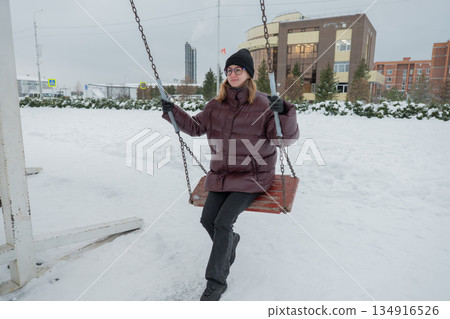 Young caucasian woman posing with one foot on swing in snowy plaza, maroon puffer and black beanie, confident stance, playful expression, modern architecture backdrop, quick urban break during outdoor 134916526