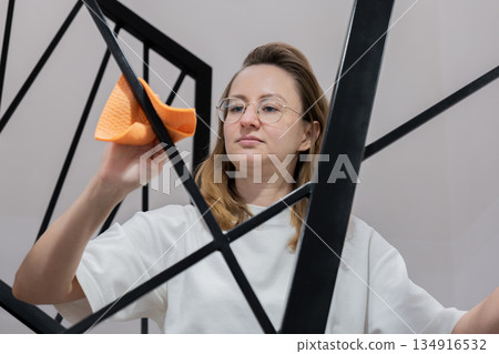 young woman cleaning glass mirror with orange cloth, wearing white shirt and glasses, focused expression, wiping black geometric frame, close-up interior scene, performing routine home maintenance young woman cleaning glass mirror with orange cloth, wearing white shirt and glasses, focused expression, wiping black geometric frame, close-up interior scene, performing routine home maintenance 134916532