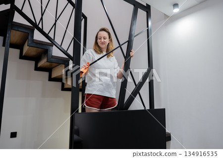 caucasian woman cleaning metal staircase railing, wearing orange glove and holding microfiber cloth, focused expression, standing near black tv console under stairs, modern minimalist caucasian woman cleaning metal staircase railing, wearing orange glove and holding microfiber cloth, focused expression, standing near black tv console under stairs, modern minimalist 134916535