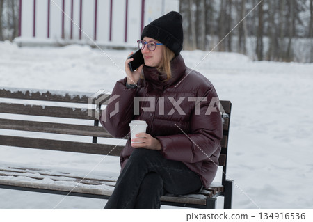 woman speaking on phone while sipping coffee, creating content vibe with natural gestures, engaged expression and relaxed posture on bench, puffer jacket and winter hat, urban outdoor scene woman speaking on phone while sipping coffee, creating content vibe with natural gestures, engaged expression and relaxed posture on bench, puffer jacket and winter hat, urban outdoor scene 134916536