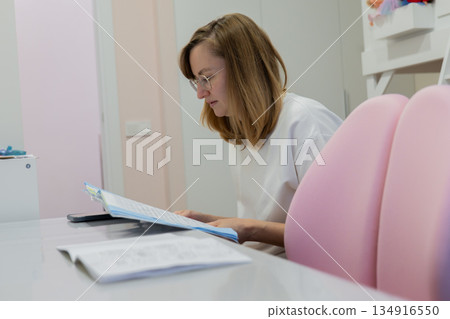 Caucasian mother grading homework at desk, wearing glasses, focused expression, organizing worksheets and notes beside pastel chairs and shelf, planning next tutoring session, calm bright domestic 134916550