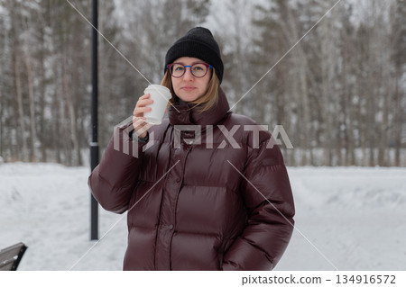 caucasian woman closeup holding latte, maroon puffer and black beanie, soft steam visible, glasses framing face, thoughtful expression, muted winter palette, cozy morning mood, calm focus and warmth 134916572