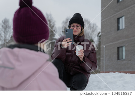 Woman kneeling filming child in snow, holding coffee and phone, child wearing purple hat playing with yellow shovel, apartment building and birch trees in background, candid urban winter family 134916587