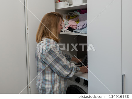 Woman loading dryer in closet, sorting soft towels into basket, wearing flannel shirt, stacked washer and dryer, open cabinet with folded linens, warm natural light, calm domestic atmosphere, Woman loading dryer in closet, sorting soft towels into basket, wearing flannel shirt, stacked washer and dryer, open cabinet with folded linens, warm natural light, calm domestic atmosphere, 134916588