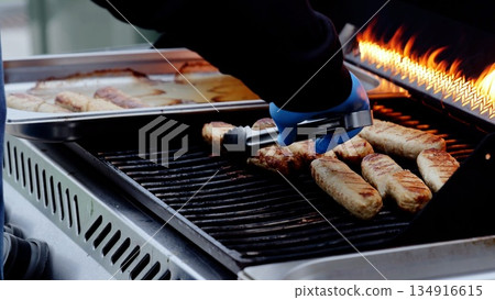 Chef wearing blue protective gloves grilling sausages on barbecue, flames flickering, cooked meat tray beside cooking surface 134916615