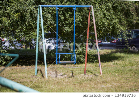 A swing set sits in a park with grass around it. The swing hangs from a metal frame with blue and red colors. Cars park in the background A swing set sits in a park with grass around it. The swing hangs from a metal frame with blue and red colors. Cars park in the background 134919736