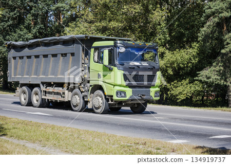 A green dump truck is driving on a road. Trees line the roadside. It is a sunny day in the countryside A green dump truck is driving on a road. Trees line the roadside. It is a sunny day in the countryside 134919737