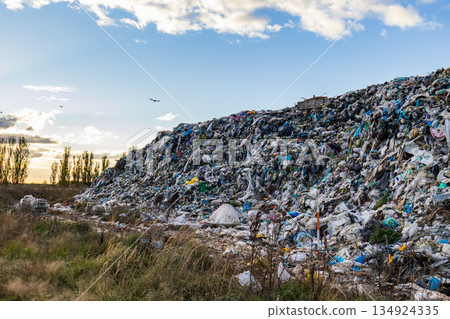 A large landfill contains heaps of garbage and waste materials under cloudy skies with some birds flying above the site 134924335