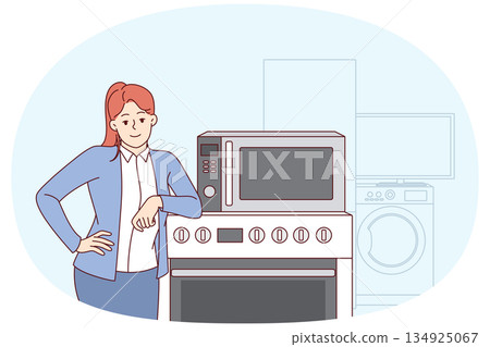 Woman selling household appliances stands near oven and washing machine and looks at camera 134925067