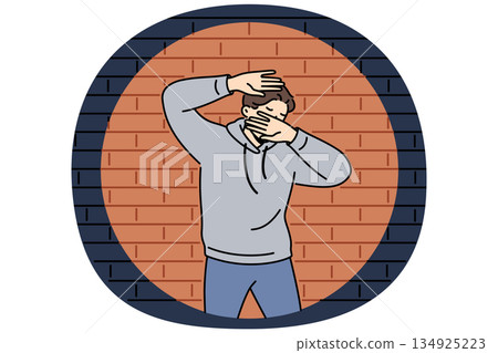 Man criminal in light of police lantern stands near brick wall and covers face with hands 134925223