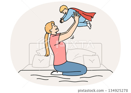 Happy mother lifts up newborn son in superhero costume, sitting on bed in bedroom 134925278