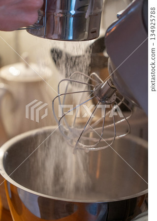 Hands sift flour into mixer bowl while preparing apple charlotte batter on wooden kitchen counter, capturing focused home baking moment in warm light 134925788