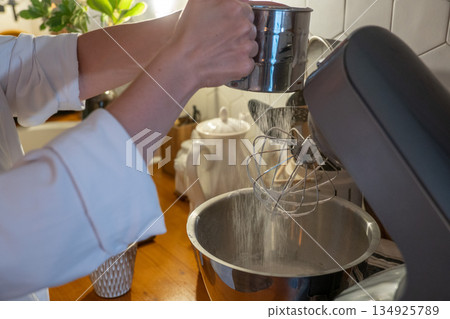 Hands sift flour into mixer bowl while preparing apple charlotte batter on wooden kitchen counter, capturing focused home baking moment in warm light 134925789