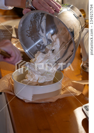 Hands pour airy apple charlotte batter from metal bowl into baking pan on kitchen counter, capturing cozy home baking moment with warm light 134925794