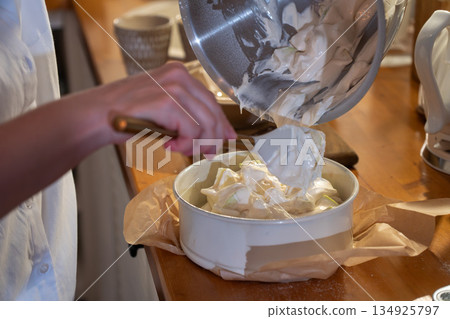 Hands pour airy apple charlotte batter from metal bowl into baking pan on kitchen counter, capturing cozy home baking moment with warm light 134925797