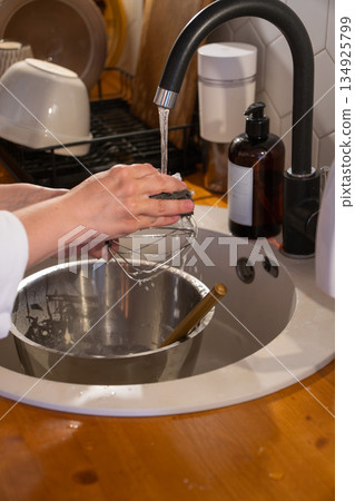 Hands rinse metal whisk over kitchen sink with mixing bowl and wooden spoon below, showing calm everyday dishwashing moment 134925799