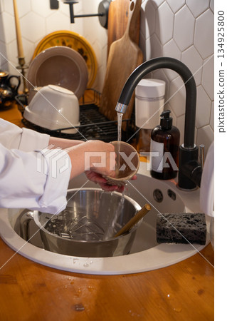 Hands rinse a ceramic cup under running water in a modern kitchen sink, with metal bowl, sponge, and soap creating a warm everyday home mood 134925800