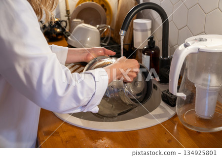 Hands wash metal mixing bowl in modern kitchen sink with running water, showing calm everyday home routine in warm indoor light Hands wash metal mixing bowl in modern kitchen sink with running water, showing calm everyday home routine in warm indoor light 134925801