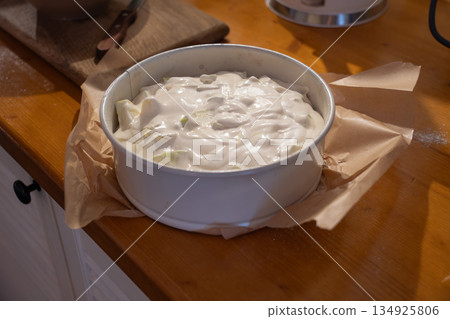 Apple charlotte batter fills round baking pan lined with parchment on wooden kitchen countertop, showing sliced apples in creamy dough 134925806