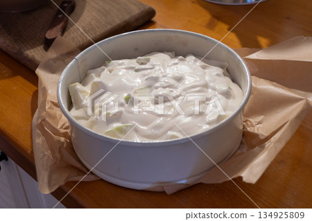 Ready apple charlotte batter rests in round baking pan lined with parchment on wooden kitchen counter, showing smooth creamy texture with apple pieces 134925809