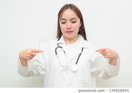 Portrait of young Asian female doctor against white background pointing finger Portrait of young Asian female doctor against white background pointing finger 134926650