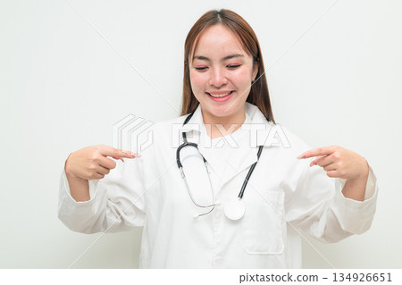 Portrait of young Asian female doctor against white background pointing finger Portrait of young Asian female doctor against white background pointing finger 134926651