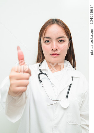 Portrait of young Asian female doctor against white background giving thumbs up gesture Portrait of young Asian female doctor against white background giving thumbs up gesture 134926654
