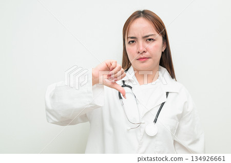 Portrait of young Asian female doctor against white background giving thumbs down gesture Portrait of young Asian female doctor against white background giving thumbs down gesture 134926661