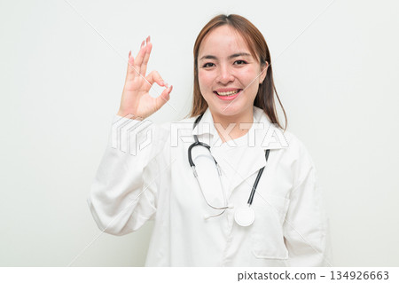 Portrait of young Asian female doctor against white background making OK sign gesture Portrait of young Asian female doctor against white background making OK sign gesture 134926663
