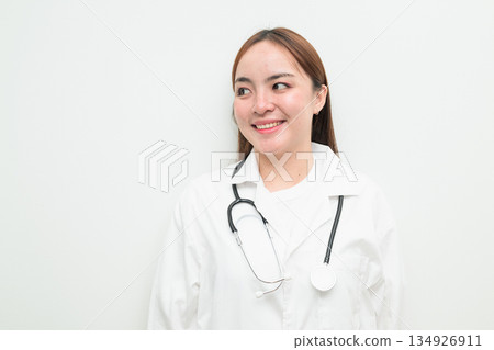Portrait of young happy Asian female doctor against white background smiling and thinking Portrait of young happy Asian female doctor against white background smiling and thinking 134926911