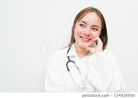 Portrait of young happy Asian female doctor against white background hand on chin thinking Portrait of young happy Asian female doctor against white background hand on chin thinking 134926930