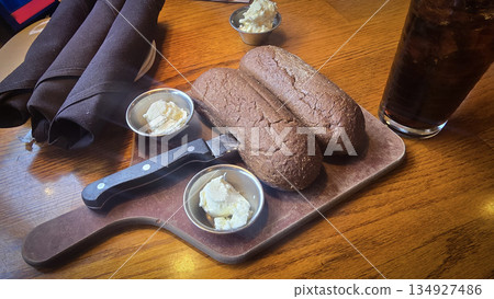 Brown Australian rye loaves rest on a rustic wooden board with small butter cups, a sharp knife, and a glass of beverage, creating a cozy, aesthetic dining scene 134927486